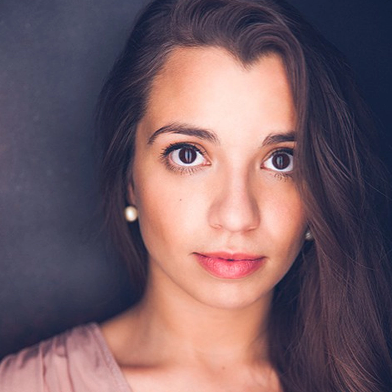 Amanda Lopez, a young woman with long brown hair and big brown eyes, looks directly at the camera. She has a neutral expression, wears pearl earrings and a pale pink top. Her hair is swept over one shoulder against a dark, softly blurred background.