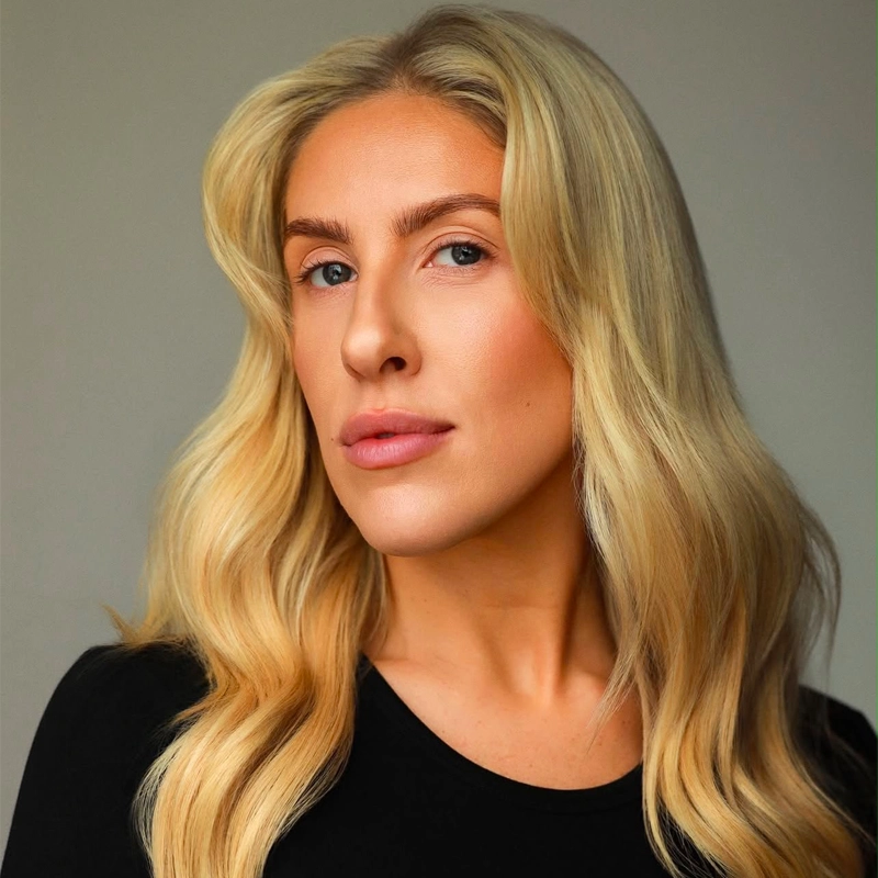 A person with long, wavy blonde hair and fair skin poses against a plain gray background, wearing a black top. Reminiscent of Natalie Paris, they have blue eyes, defined brows, and a neutral facial expression, looking confidently at the camera.
