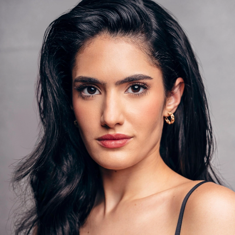 A woman with long, wavy black hair and fair skin looks confidently at the camera. She has defined eyebrows, subtle makeup with pink lipstick, and wears gold hoop earrings and a black spaghetti strap top. The background is neutral gray.