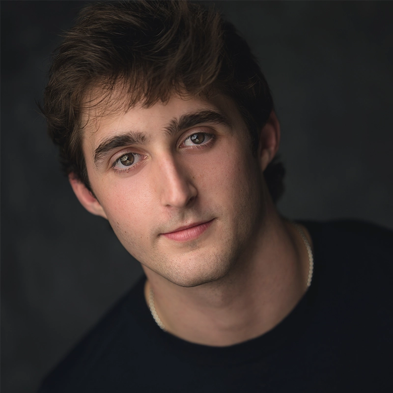 A young man with wavy brown hair and fair skin looks at the camera with a slight, relaxed smile. He wears a dark shirt and a thin chain necklace. The background is dark and softly blurred, highlighting his face and expression.