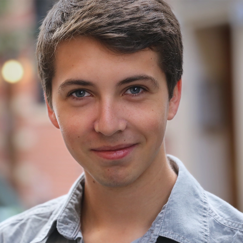 Jarrod Allan, a young person with short brown hair and blue eyes, smiles slightly at the camera. They are wearing a light gray collared shirt and standing outdoors, with a blurred background of warm colors and soft lighting.