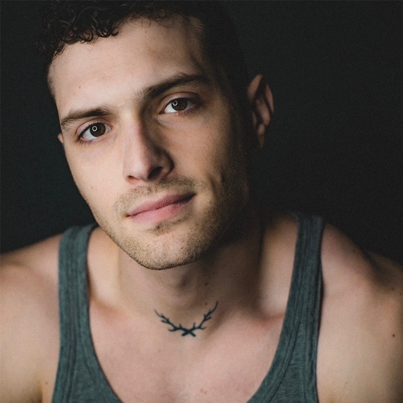 A young man with short curly hair and light skin wears a gray tank top. He faces the camera with a slight smile and gentle expression. A small black antler tattoo is visible at the base of his neck. The background is dark and plain.