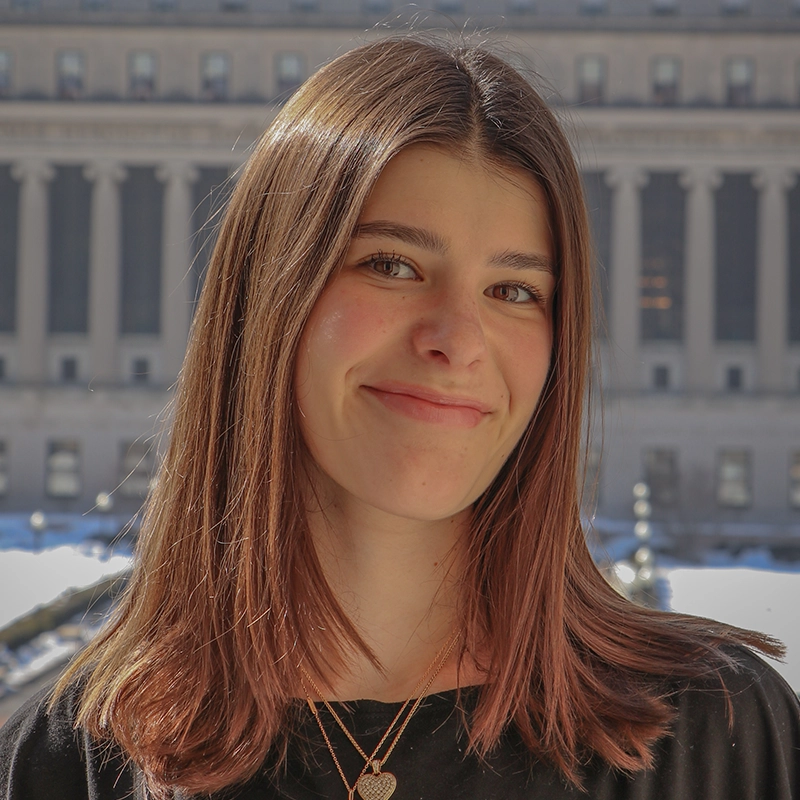 A young woman with straight, shoulder-length brown hair smiles softly. She wears a black top and layered gold necklaces. The background shows a large, sunlit building with columns and rows of windows, suggesting an outdoor urban setting.