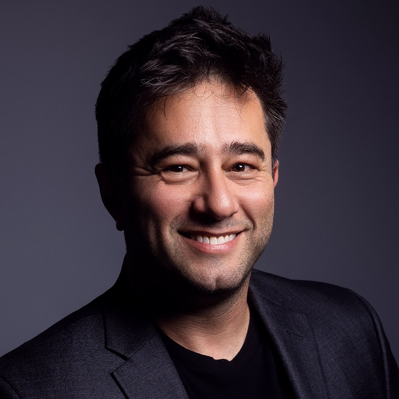 A man with dark hair and stubble smiles at the camera. He is wearing a dark blazer over a black shirt. The background is a plain, dark gradient. The lighting highlights his face, creating a professional studio portrait effect.