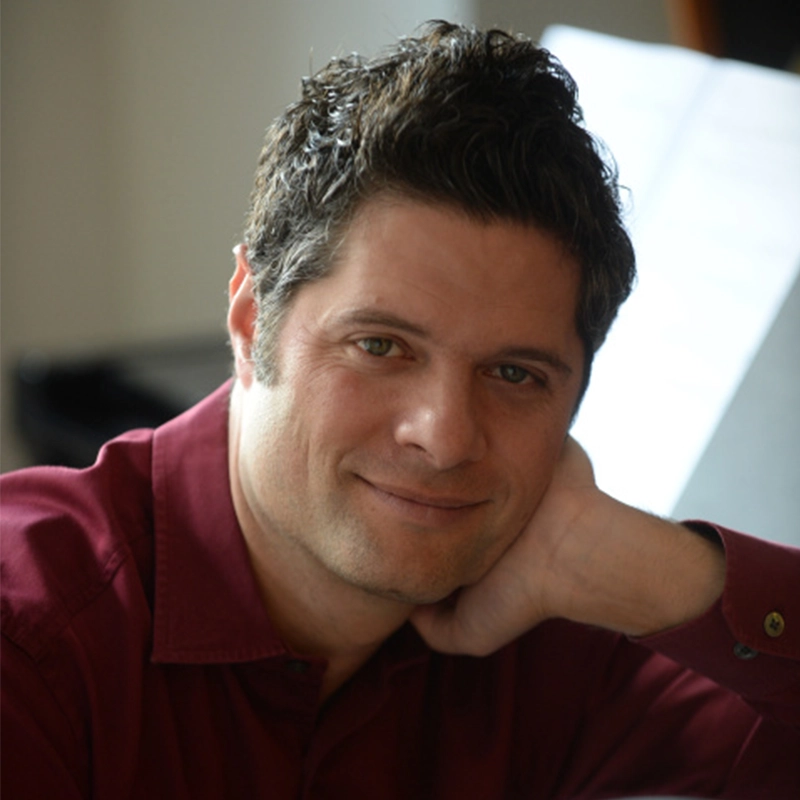A man with short, dark hair and light skin smiles gently at the camera. He is wearing a maroon button-up shirt and rests his head on his left hand. The background is softly blurred, showing what appears to be a piano and sheet music.
