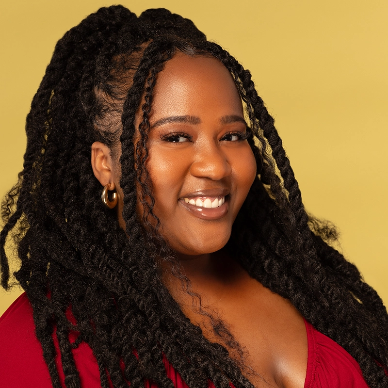 A smiling woman with long, thick twists in her hair—Aneesa Folds—wears gold hoop earrings and a deep red top. She poses against a solid yellow background, looking directly at the camera with a warm, confident expression.