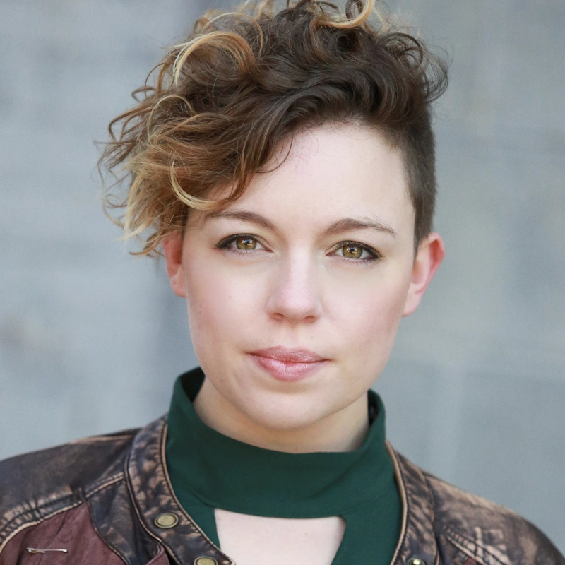 A person with short, curly hair—brown with blonde highlights—styled to one side, looks directly at the camera. Hannah MacDonald wears a green high-collared top and a brown, distressed leather jacket. The blurred gray background suggests an outdoor wall.