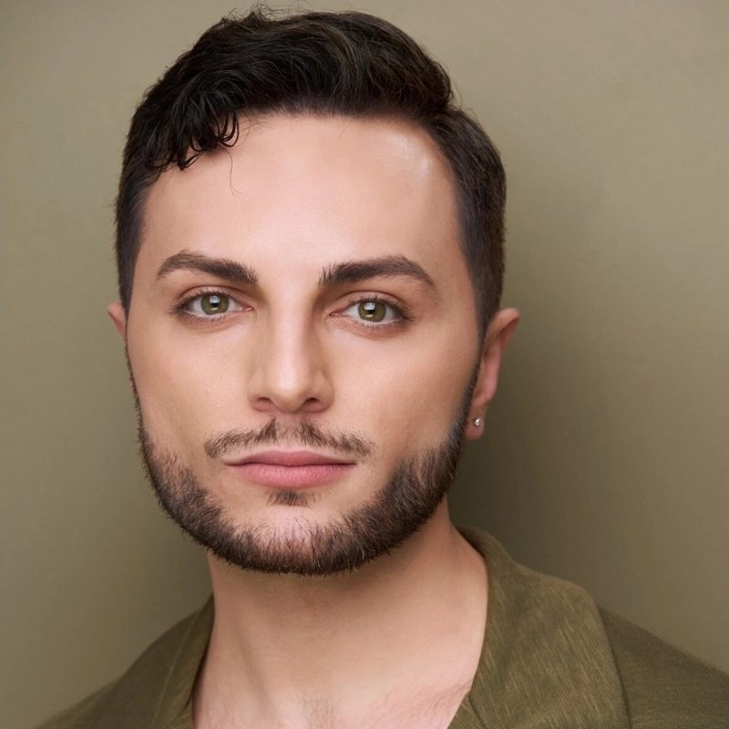 A young man with short dark hair, light skin, hazel eyes, and a trimmed beard looks at the camera. He wears a green shirt and a small earring in his left ear. The background is plain beige, creating a simple yet polished portrait reminiscent of Joseph C. Townsend’s understated elegance.