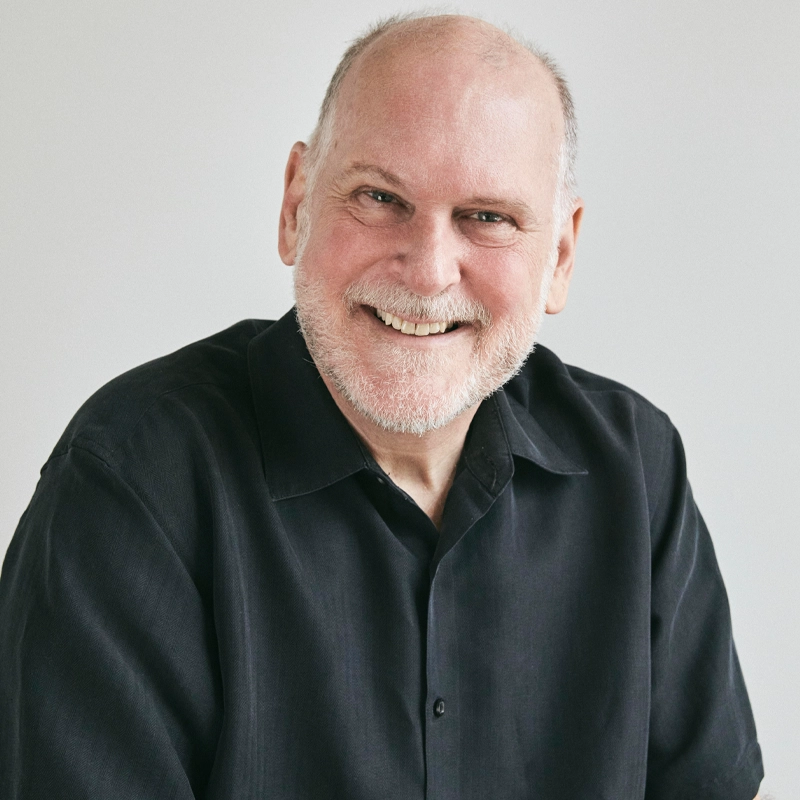 A smiling older man with a bald head and white beard wears a black button-up shirt. He faces the camera against a plain, light grey background. His relaxed posture and expression convey warmth and friendliness.