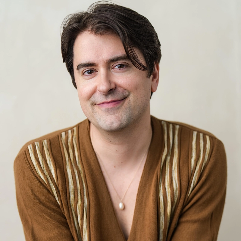 A person with fair skin and dark brown, side-parted hair smiles gently at the camera. They wear a brown sweater with light vertical stripes and a pearl necklace. The background is a light, neutral color.