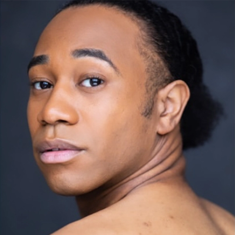 A person with medium brown skin and dark hair pulled back looks over their shoulder towards the camera, with a neutral expression and soft lighting against a dark background.