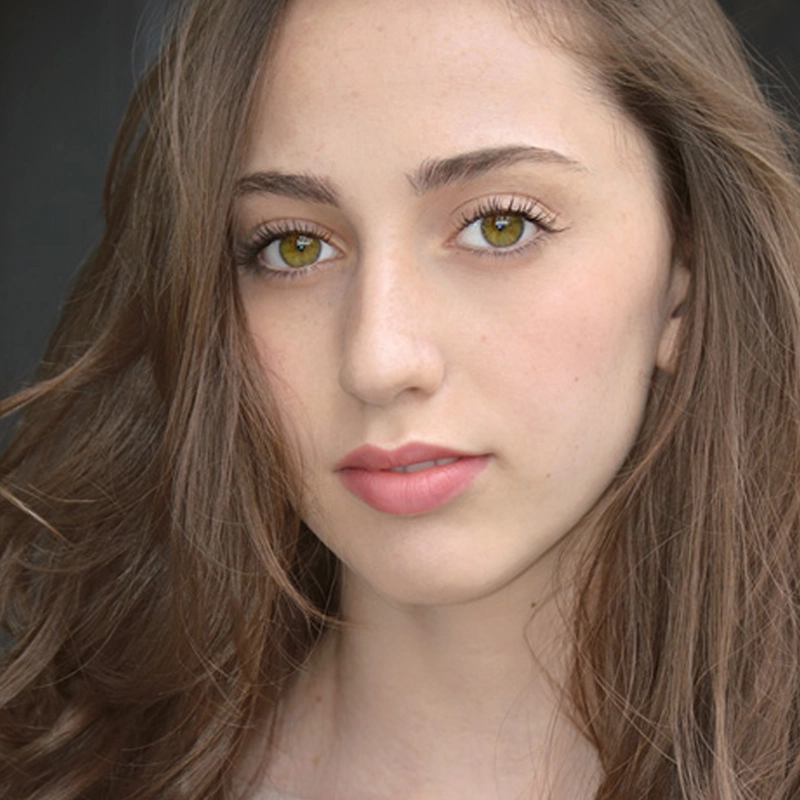 A young woman with light skin, long brown hair, and hazel eyes gazes softly at the camera. Laura Sky Herman has natural makeup, with defined eyebrows and pink lips, and is posed in front of a dark, neutral background. Her hair flows gently around her face.