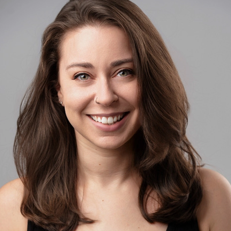 Jessie Linden, a young woman with long brown hair and light skin, smiles at the camera. She has a small nose ring and is wearing a black sleeveless top. The plain light gray background adds a professional, studio portrait feel.