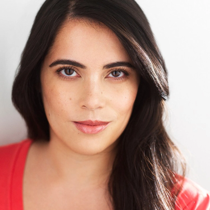 Chrissy Pardo, a woman with long, dark hair and fair skin, looks directly at the camera. She has brown eyes, lightly arched eyebrows, and wears a red top. The white, softly lit background highlights her face and gentle expression.