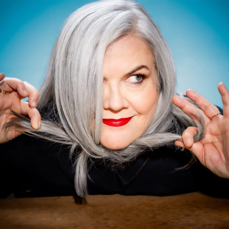 A woman with long gray hair, red lipstick, and a playful expression holds her hair across her upper lip like a mustache. She wears a black top and poses in front of a bright blue background. Her eyes glance sideways, and she smiles mischievously.