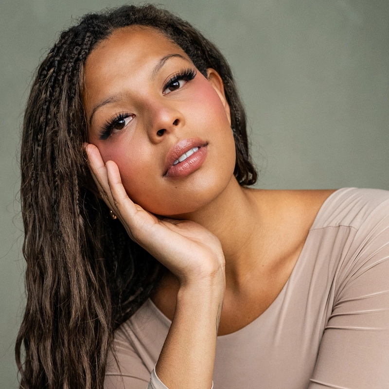 A young woman with long, braided brown hair rests her head on her hand. She has glowing skin, natural makeup with rosy cheeks and glossy lips, and wears a beige top. The background is a soft, muted green. She gazes calmly at the camera.