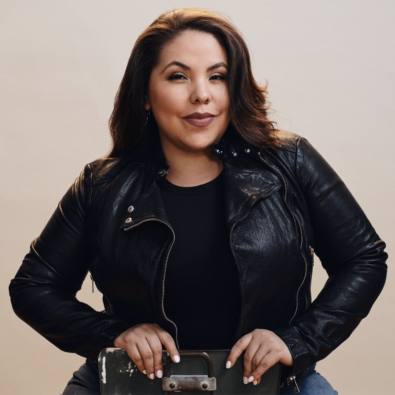 Florencia Cuenca, with medium-length wavy brown hair, sits facing the camera in a black leather jacket and shirt, smiling slightly as she rests her hands on the back of a vintage metal chair against a plain beige background.
