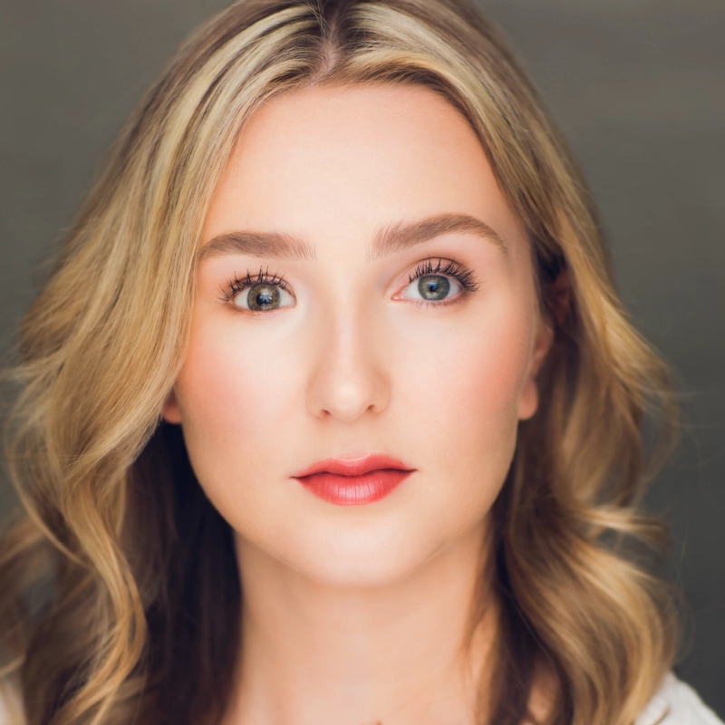Noelle Cornelius, a young woman with wavy blonde hair, fair skin, and green eyes, looks directly at the camera. She wears natural makeup with defined brows, subtle eyeshadow, mascara, and reddish-pink lipstick against a softly blurred background.