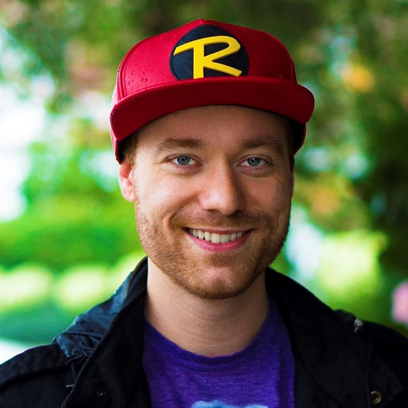 A smiling man, Justin Gomlak Greer, with a reddish beard wears a red cap featuring a yellow “R” logo in a black circle. He has blue eyes and is dressed in a purple T-shirt and dark jacket. The blurred background includes greenery and soft daylight.