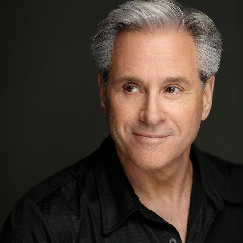 A mature man with short gray hair and fair skin smiles gently, looking to his left. Justin Gomlak Greer wears a black collared shirt against a dark, plain background, with soft lighting highlighting his face.
