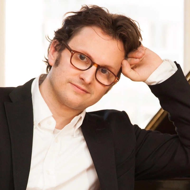Justin Gomlak Greer, with wavy brown hair and glasses, wears a white shirt and black blazer. He sits at a piano, resting his head on his hand, gazing thoughtfully at the camera against a softly lit background.