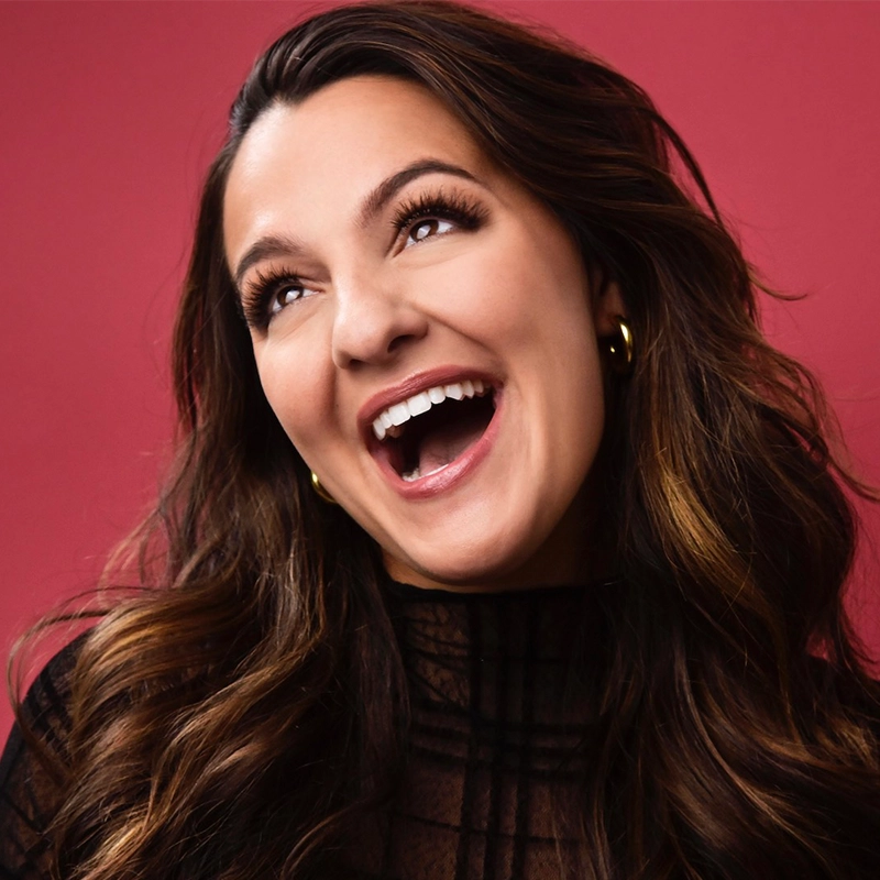 Deanna Giulietti, with long, wavy brown hair and hoop earrings, smiles widely as she looks upward. She wears a black, semi-sheer high-neck top against a deep pink background for a vibrant and joyful atmosphere.