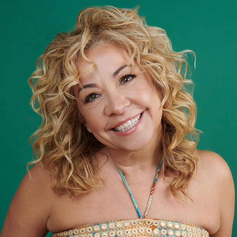 Joyce Chittick, a woman with curly, blonde hair, smiles warmly at the camera. She wears a strapless dress with a patterned design and thin halter strap. The background is solid green, and her expression is cheerful and friendly.