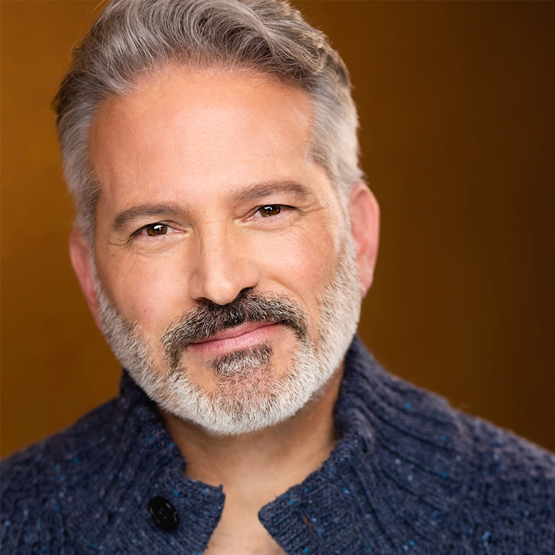 Portrait of Justin Gomlak Greer, a middle-aged man with fair skin, short gray hair, and a full salt-and-pepper beard. He wears a dark blue textured sweater with a high collar, smiling softly against a warm brown background.