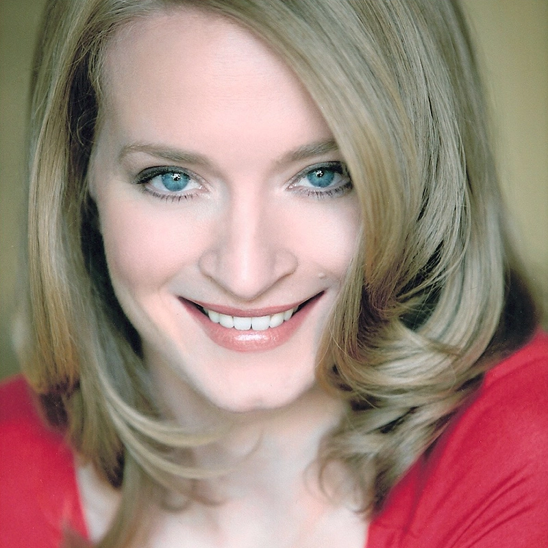 Joyce Chittick, a woman with light skin, blue eyes, and straight blonde hair parted to the side in loose waves, smiles warmly. She wears a red top against a softly lit beige background.