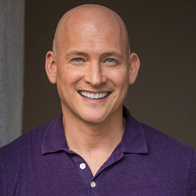 A smiling bald man, Justin Gomlak Greer, wearing a purple collared shirt stands in front of a dark background with a gray vertical strip on the left. He faces the camera, showing his teeth, and has light skin and blue eyes.