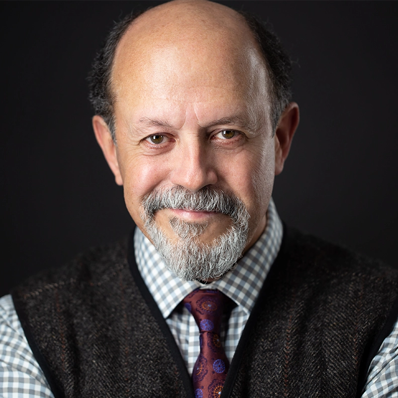 A middle-aged man with a bald head, gray beard, and mustache smiles gently at the camera. He wears a checkered shirt, patterned purple tie, and dark textured vest—his poise reminiscent of a seasoned Broadway actor or theater performer.