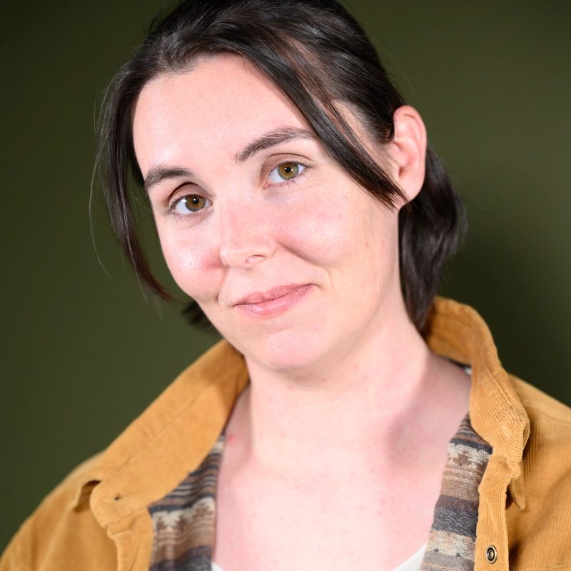 A person with fair skin, straight brown hair in a bob, and hazel eyes smiles softly. They wear a mustard yellow jacket over a patterned shirt. The background is plain olive green. The lighting is soft, highlighting their facial features.