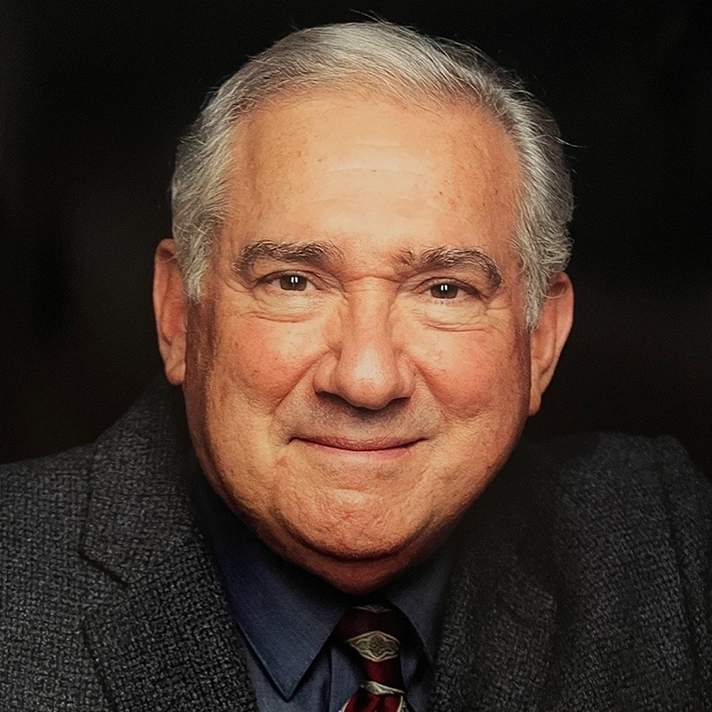 An older man with gray hair smiles warmly. He wears a dark gray textured suit jacket, a blue dress shirt, and a patterned tie. The black background draws attention to his friendly expression—a style reminiscent of Joyce Chittick's classic elegance.