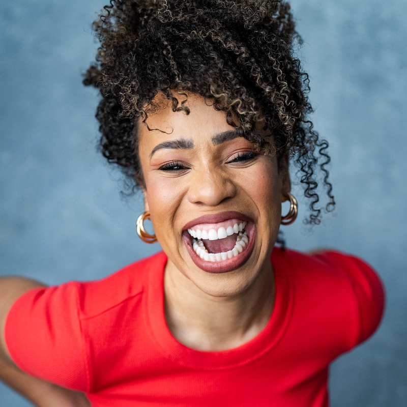 Salisha Thomas, with her curly hair, gold hoop earrings, and bright red top, smiles broadly at the camera against a soft blue background. Her relaxed posture and radiant expression exude confidence, joy, and enthusiasm.