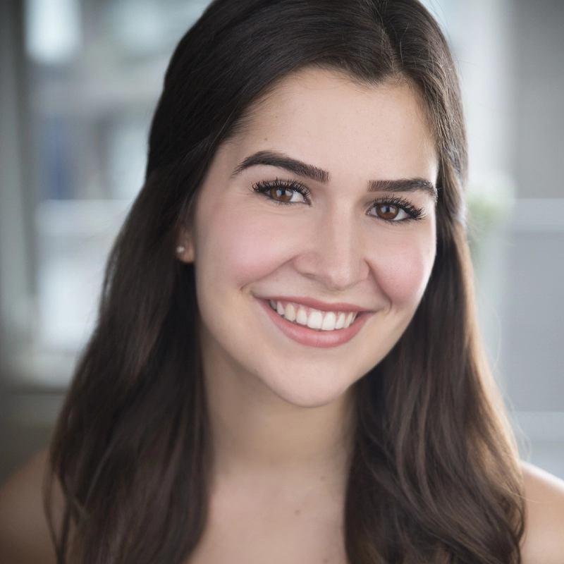 A young woman with long dark hair and brown eyes smiles at the camera. She is wearing minimal makeup and a natural lip color. The background is softly blurred with cool tones, suggesting an indoor setting with natural light. This candid portrait of Elizabeth Bedley captures her fresh and approachable charm.