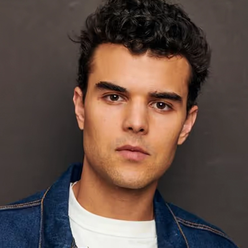 Frankie J. Gonzalez, a young man with short, curly dark hair and fair skin, stares directly at the camera. He wears a blue denim jacket over a white crew-neck shirt against a plain dark background that highlights his neutral expression.