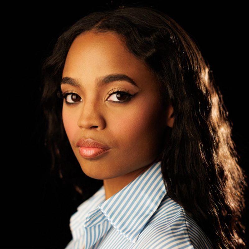Morgan Dudley, a woman with wavy black hair and smooth skin, looks confidently at the camera. She wears a blue and white striped collared shirt. Her makeup is subtle, featuring winged eyeliner and natural lipstick, with a dark background highlighting her face.