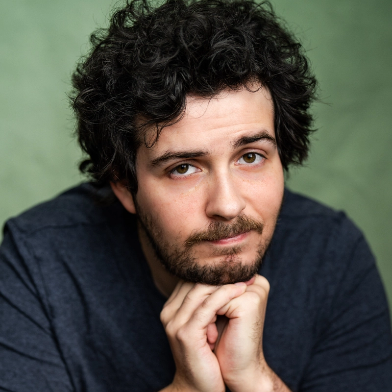 A man with curly dark hair and a short beard, resembling Asher Muldoon, rests his chin on his hands and looks at the camera with a gentle smile. He wears a dark gray shirt and sits in front of a muted green background.