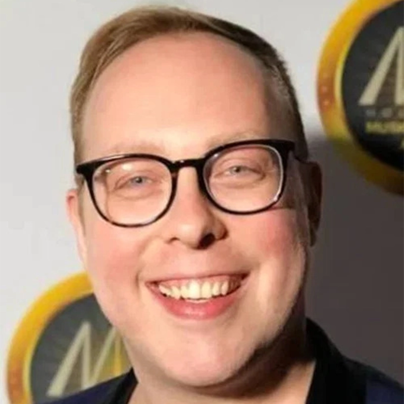 Shira Averbuch, a smiling person with light skin and short hair, wears black-rimmed glasses in front of a backdrop featuring circular gold logos.