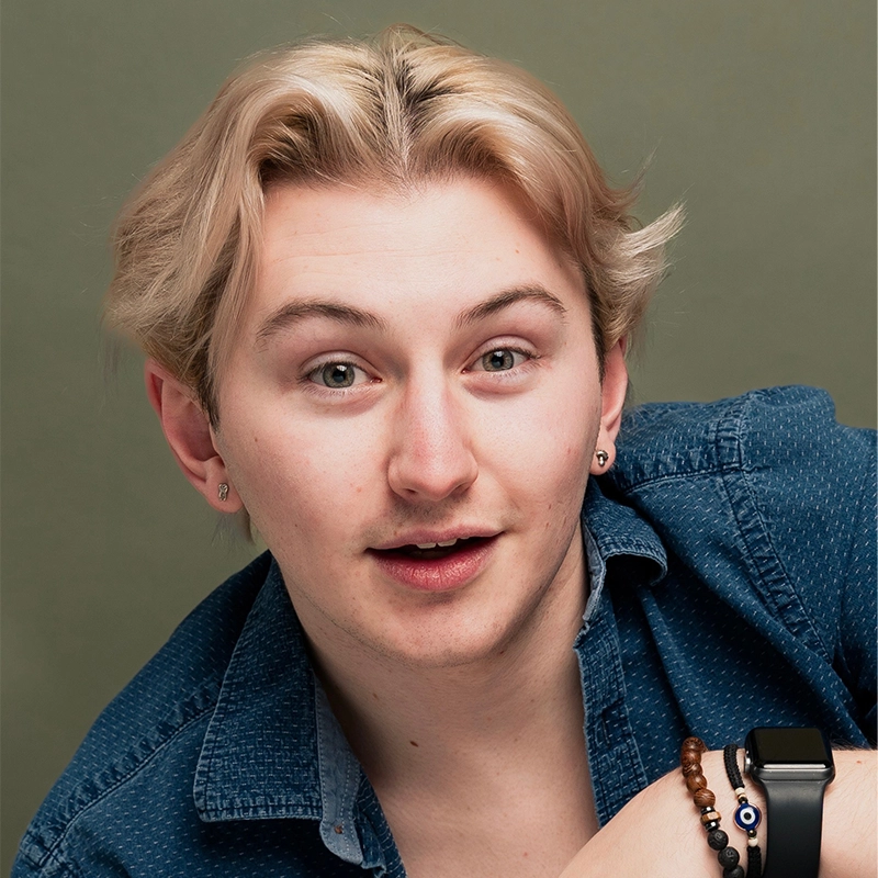 A young person with light skin and layered blond hair, Andi Maroney, gazes at the camera with a surprised expression. They wear a blue denim shirt, small hoop earrings, a smartwatch, and beaded bracelets against an olive-green background.