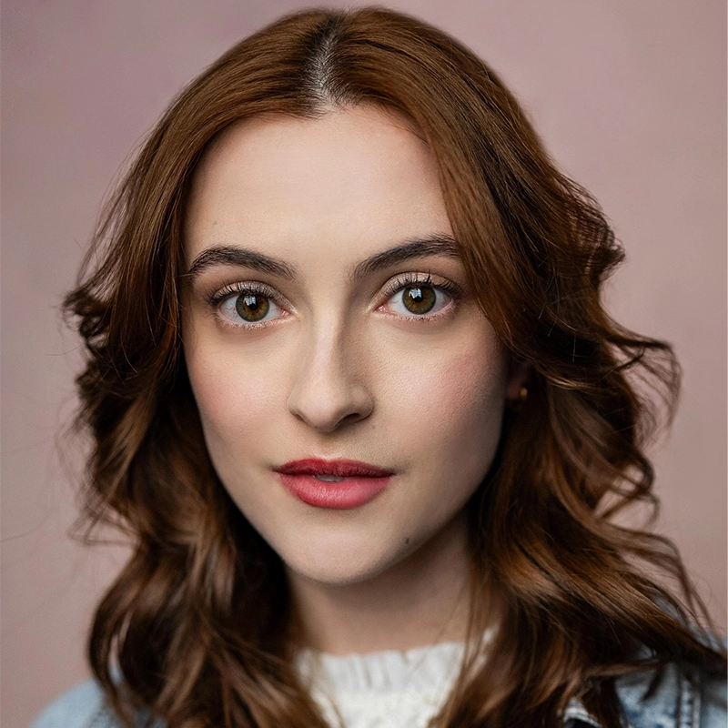 Headshot of Shira Averbuch with long, wavy auburn hair looking directly at the camera. She has light skin, brown eyes, and is wearing a denim jacket over a white top.
