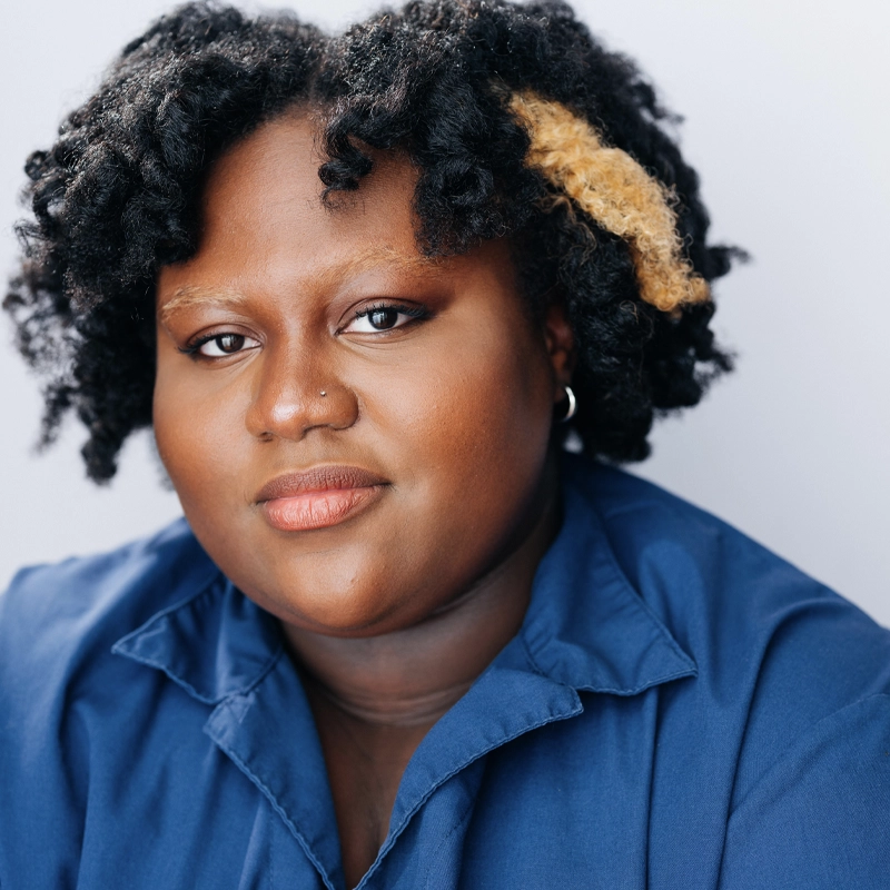 Ayana François, with deep brown skin and textured, short black hair featuring a blonde streak, smiles softly. She wears a blue collared shirt, hoop earrings, and natural makeup, posing against a plain light background.