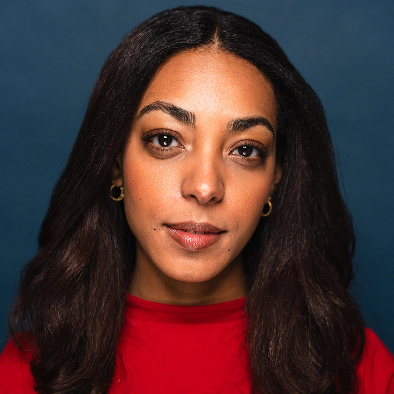 Gabriella Joy Rodriguez, with medium skin tone and long, dark wavy hair, wears a red top and small gold hoop earrings. She has brown eyes and natural makeup, looking directly at the camera with a neutral expression against a dark blue background.