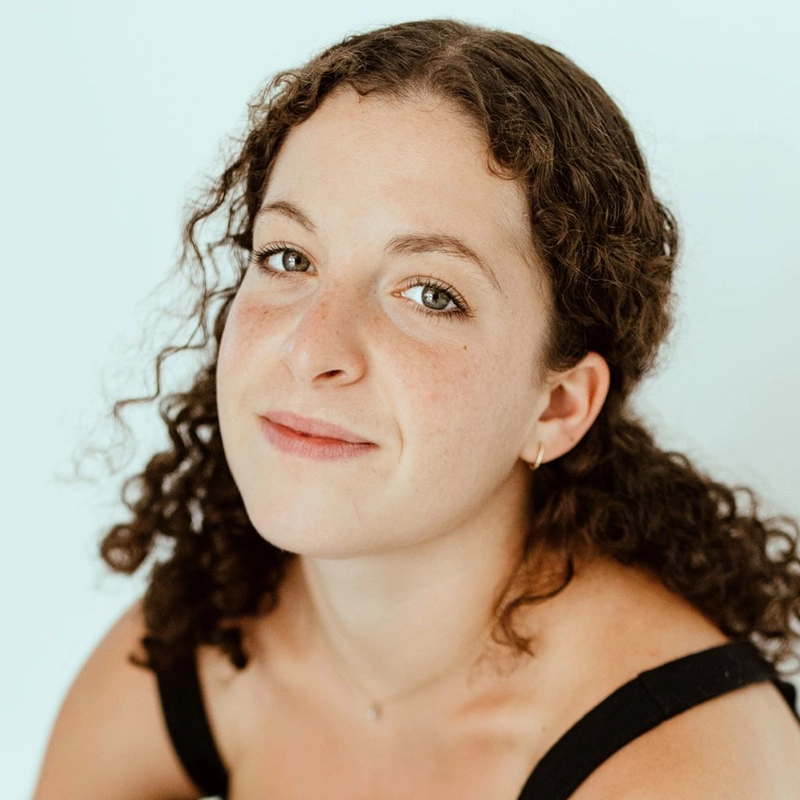 Shira Averbuch, a woman with shoulder-length curly hair, looks directly at the camera. She is wearing a black tank top against a plain light background.