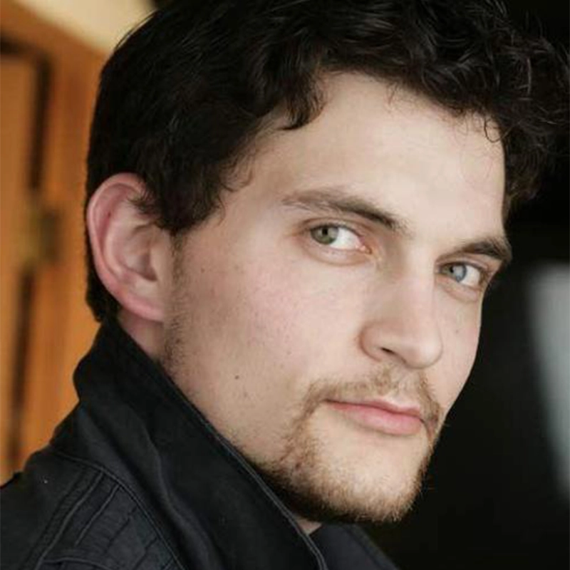 A close-up portrait of Shira Averbuch, a young man with dark, curly hair and light eyes. He has a light beard and is wearing a dark collared jacket.