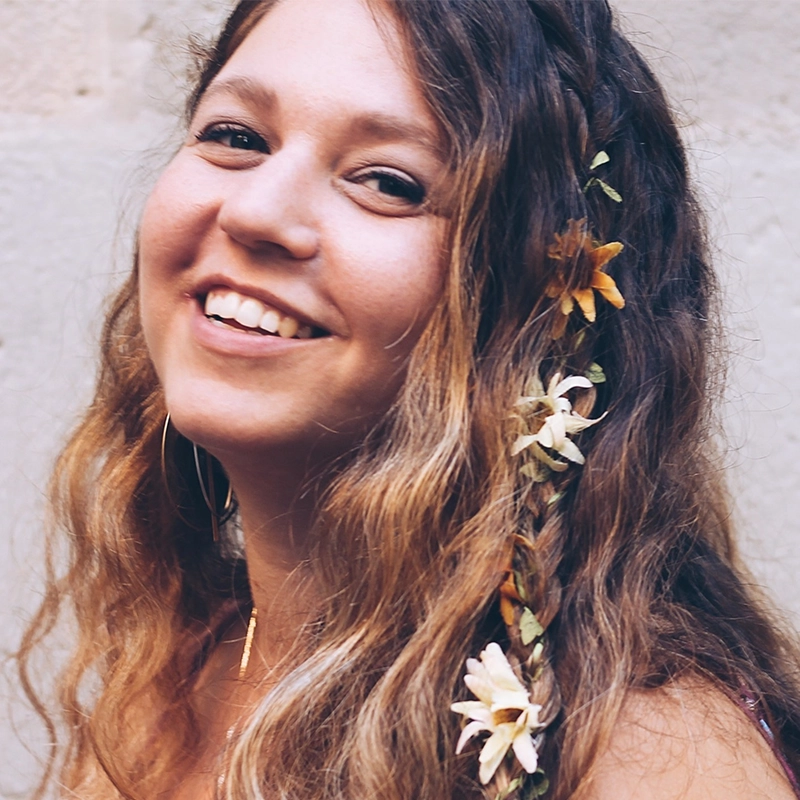 Shira Averbuch is a smiling woman with long, wavy brown hair, featuring small flowers woven into a braid along the side.
