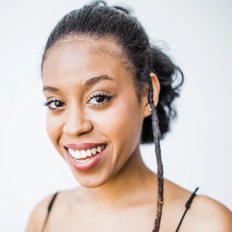 A young woman with dark skin and curly hair styled in a loose updo with one long twisted lock. She has brown eyes, is smiling brightly, and wears a black spaghetti strap top. The background is plain and white, emphasizing her face.