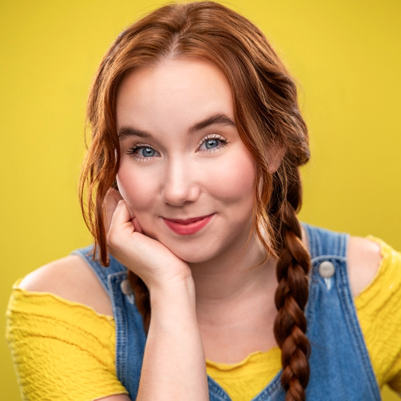 A young woman with fair skin and long red hair in two braids smiles gently, resting her chin on her hand. Lauren wears a yellow top with cut-out shoulders and blue denim overalls. The background is solid yellow, giving a bright, cheerful feel to the image.