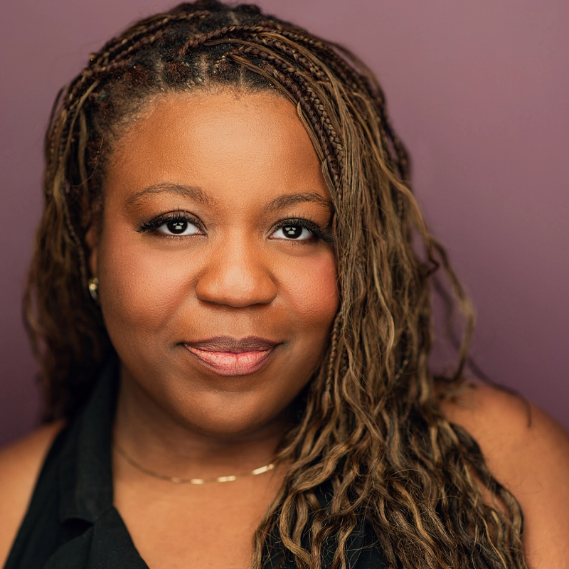 Trèjah Bostic, with long, braided brown hair, smiles softly at the camera. She wears a sleeveless black top, subtle makeup, a gold necklace, and small earrings. The solid mauve background creates a warm and professional portrait.