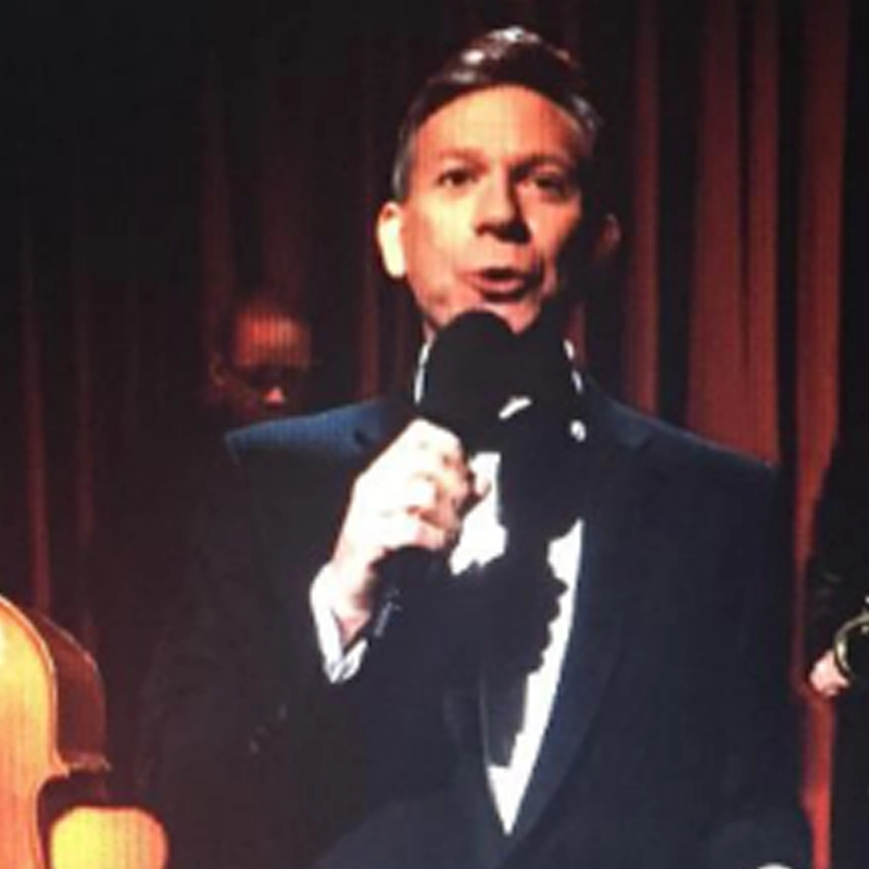 Pete Caldera, dressed in a black tuxedo, holds a microphone and speaks onstage. Behind him, musicians and a double bass are partially visible against dark curtains, suggesting a formal event or concert setting.
